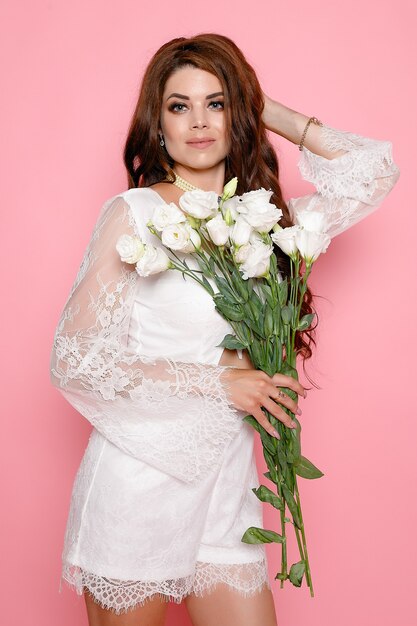 Beautiful girl isolated on pink background. Standing, looking at camera. White dress and shoes. Beautiful hairstyle and makeup. holding spring pink flower bouquet. Magnificent hair.