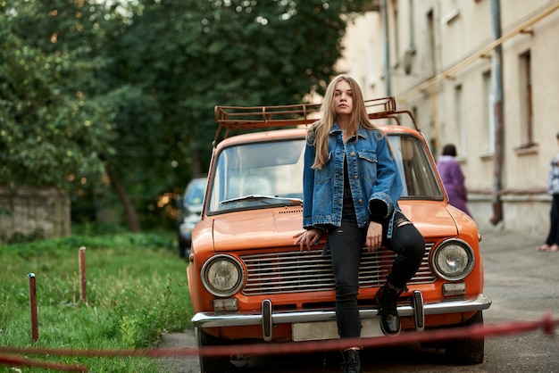 ジーンズとジャケット、オレンジ色のレトロな車のボンネットの上に座っている美しい少女。
