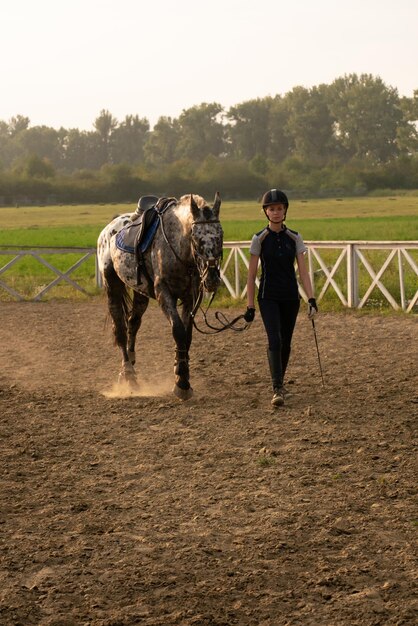 空と緑の野原に特別な制服を着た美しい少女騎手が馬の横に立っています...