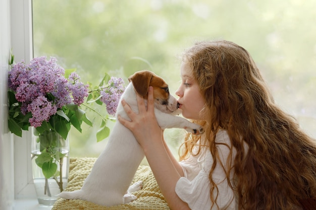 Beautiful girl kisses her puppy in nose on the window. Friendship, care, happiness concept.