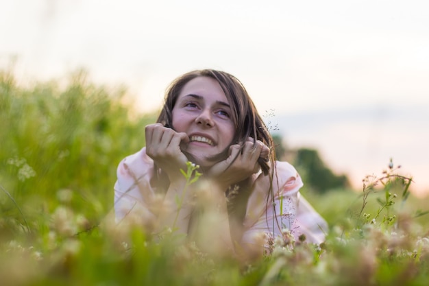 A beautiful girl lies in the grass and wildflowers holding head on hands and smiles.