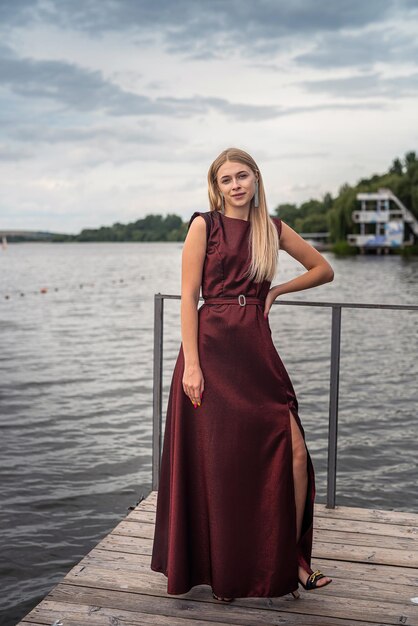 Красивая девушка в длинном модном темно-красном платье возле пруда в городском парке