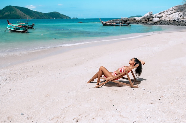 赤と白のストライプのビキニのラウンジャーで美しい少女。ビーチでリラックスしたアジアのブルネット。熱帯の休暇。タイのボートと背景に青い海