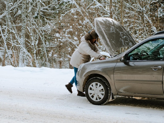 冬の雪道で壊れた車のエンジンの近くの美しい少女。凍結して助けを求める