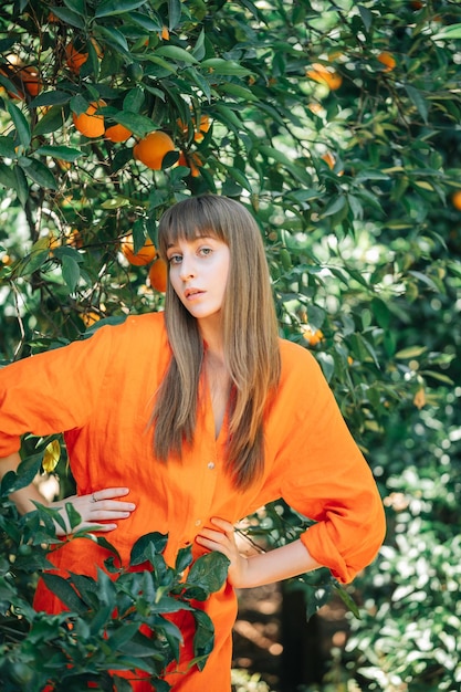 La bella ragazza in vestito arancione sta posando alla macchina fotografica mettendo le mani sulla vita nel giardino d'arancio