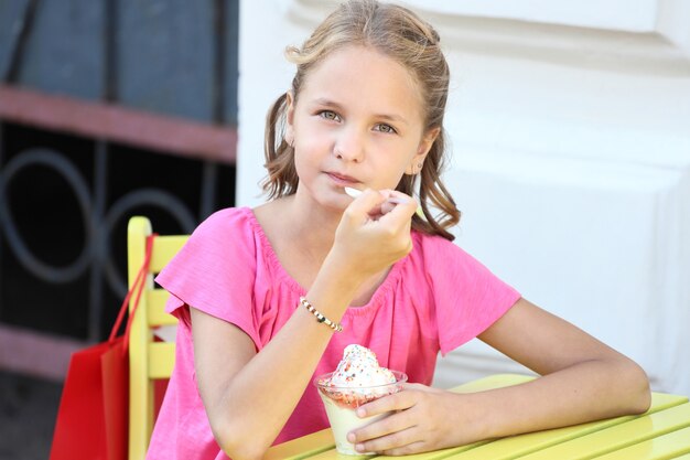 Красивая девушка в розовой футболке сидит за желтым столом и ест мороженое