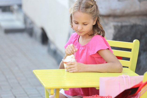 Beautiful girl in a pink t-shirt sits at a yellow table and eats ice cream