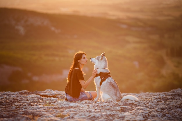 Bella ragazza gioca con un cane, husky grigio e bianco, in montagna al tramonto. ragazza indiana e il suo lupo