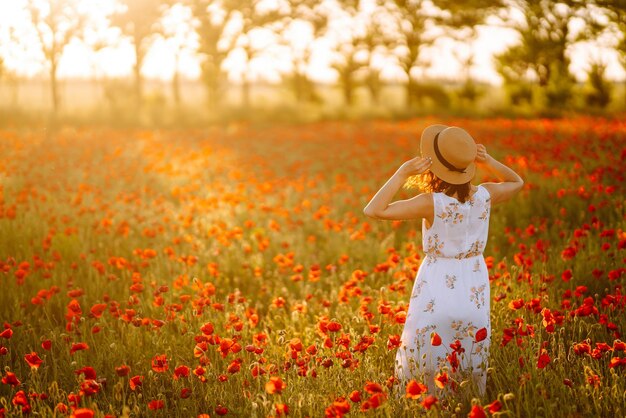 Красивая девушка на поле мака на закате в белом платье и шляпе