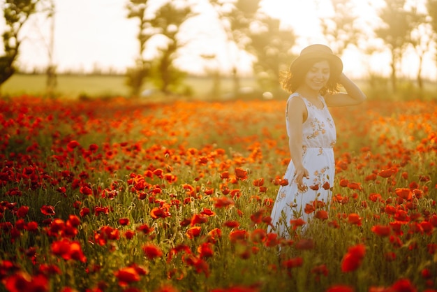 Beautiful girl in the poppy field at sunset in a white dress and hat