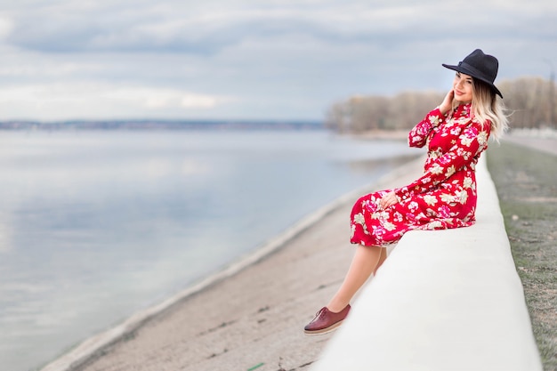 Beautiful girl in a red dress and black hat sits on the waterfront