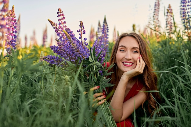 Beautiful girl in a red dress with a bouquet of lupine flowers in the field