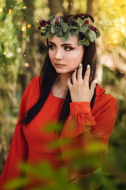 A beautiful girl in a red dress, with long dark hair and a flower wreath on her head walks through the forest.