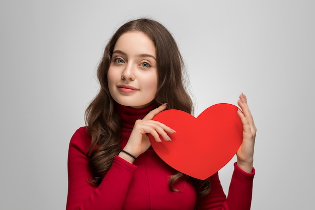 Beautiful girl in a red sweater is holding red cardboard heart She looking at the camera smiling