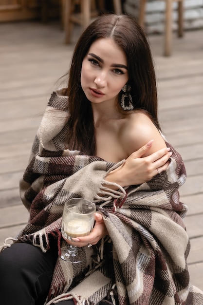 Beautiful girl resting and drinking coffee sitting in cafe on the terrace, wrapped in a woolen plaid blanket. Autumn morning. Beautiful young woman with cup of coffee outdoors on autumn day