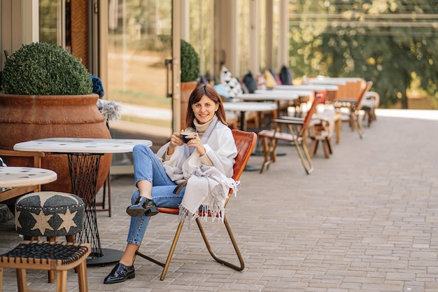 Photo beautiful girl resting and drinking coffee sitting  in cafe on the terrace, wrapped in a woolen plaid blanket. autumn morning.