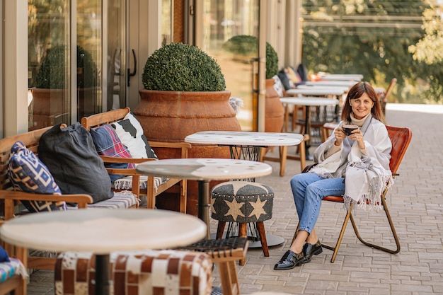 Beautiful girl resting and drinking coffee sitting  in cafe on the terrace, wrapped in a woolen plaid blanket. Autumn morning.