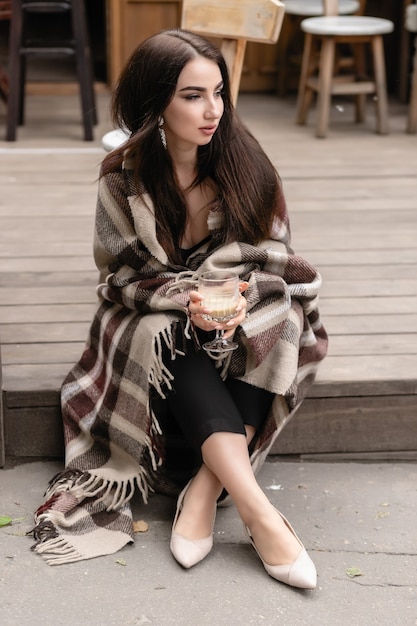 ウールの格子縞の毛布に包まれて、テラスのカフェに座って休んでコーヒーを飲む美しい少女。