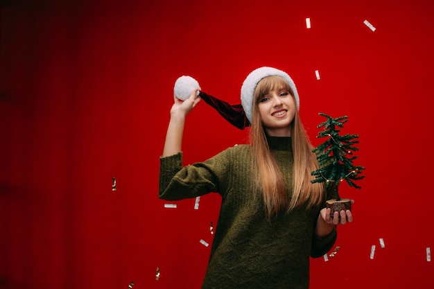 Красивая девушка в новогодней шапке держит в руках маленькую елку на красном фоне