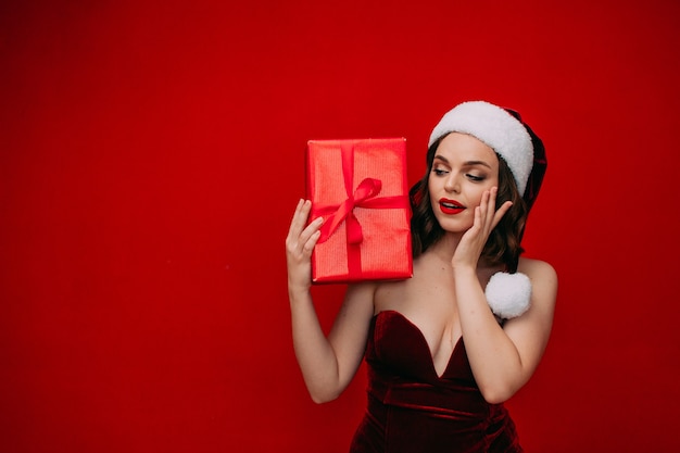 A beautiful girl in a Santa hat holds a New Years gift in her hands on a red background
