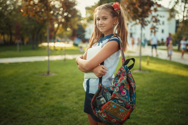 学齢期の美しい少女が木に立ってタブレットを持っています。スカートと靴のシャツを着ている女子高生