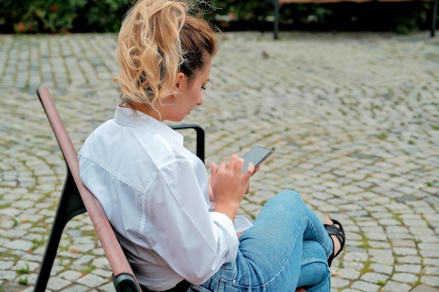 A beautiful girl sits on a bench and holds a phone in her hands the girl orders food through the phone the girl is talking on the phone laughing happy surprised