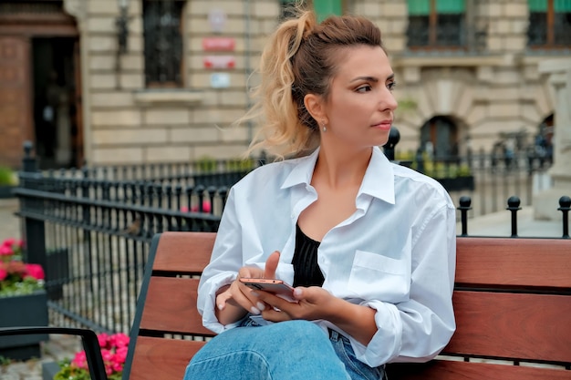 A beautiful girl sits on a bench and holds a phone in her hands the girl orders food through the phone the girl is talking on the phone laughing happy surprised