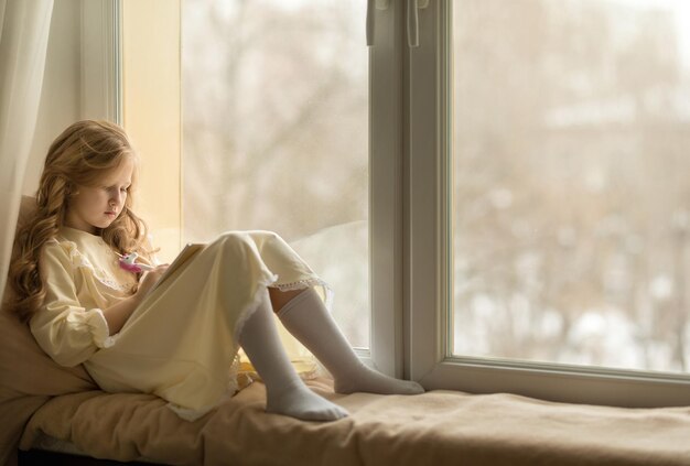 Beautiful girl sits on the windowsill and writes in a notebook