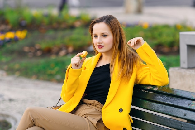 Beautiful girl sitting on a bench and holding yellow macaroon in