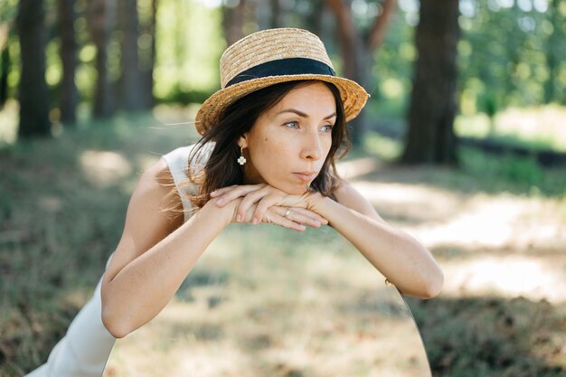 Beautiful girl in small straw hat and in white suit with big mirror in the big public park