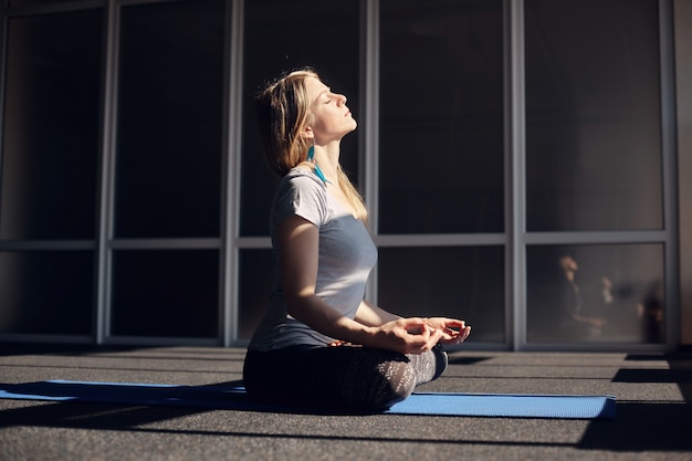 A beautiful girl in sports clothes meditates during yoga classes. The blonde is sitting in the Lotus position, relaxing, exposing her face to the sun. The concept of a healthy lifestyle. Side view