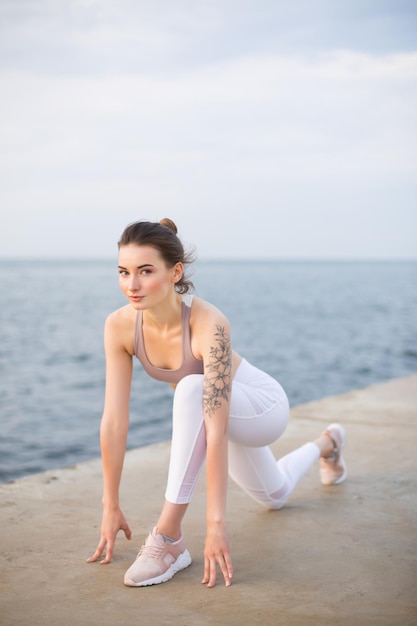 Beautiful girl in sporty top and white leggings practicing yoga with sea view on background. Young woman dreamily looking in camera while stretching by the sea