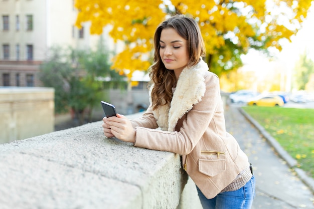 Beautiful girl standing  autumn day Holds mobile phone in hand looks at the screen.