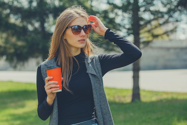 Красивая девушка стоя на улице с кофе и очками.