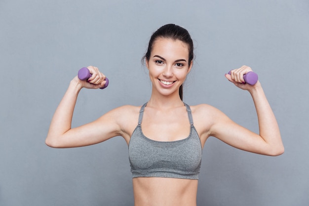 Beautiful girl standing with dumbbells and smiling isolated on grey background