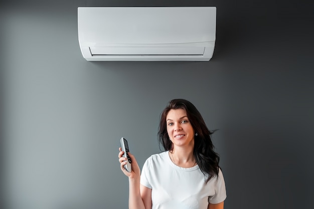 A beautiful girl stands under air conditioning and a stream of fresh cold air against a gray wall. The concept of heat, cool air, cooling, freshness.