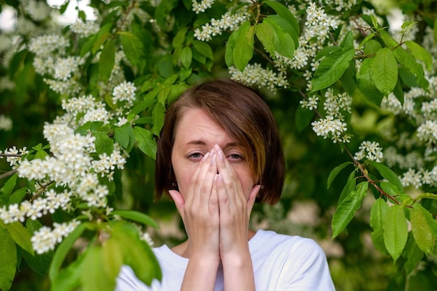 A beautiful girl stands by the blossoming bird cherry and sneezes She suffers from an allergy to flowering in the spring