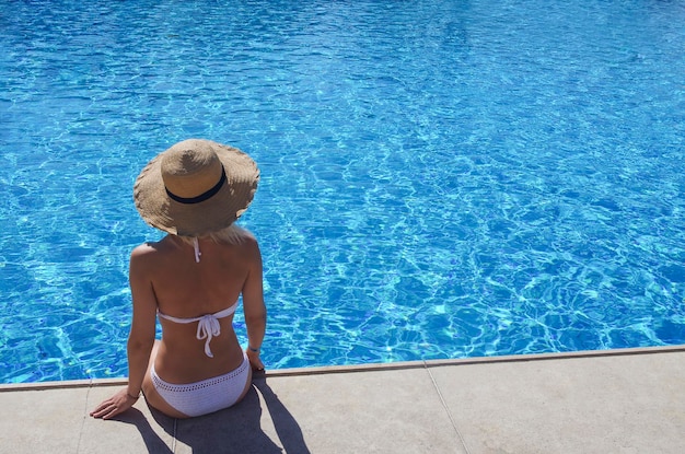 Beautiful girl in a straw hat sits on the edge of a large swimming pool Girl on the background of the pool