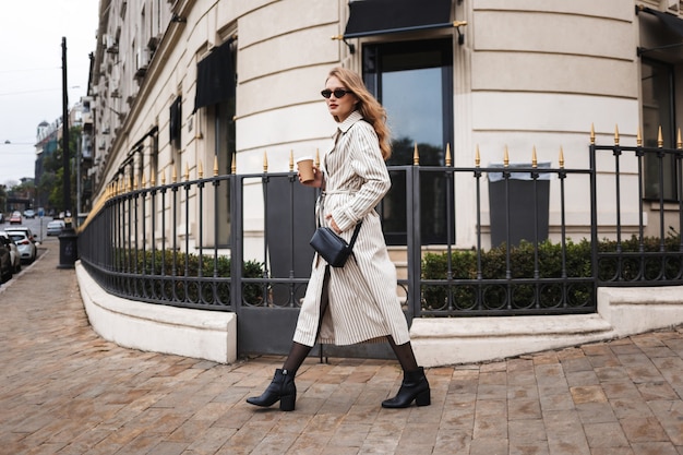 Beautiful girl in striped trench coat and sunglasses with little black cross bag holding coffee to go in hand while thoughtfully walking around cozy city street