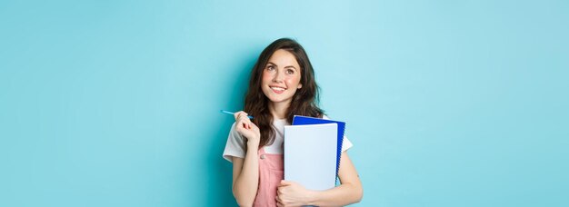 Beautiful girl student looking thoughtful at top copy space standing with notebooks homework and person