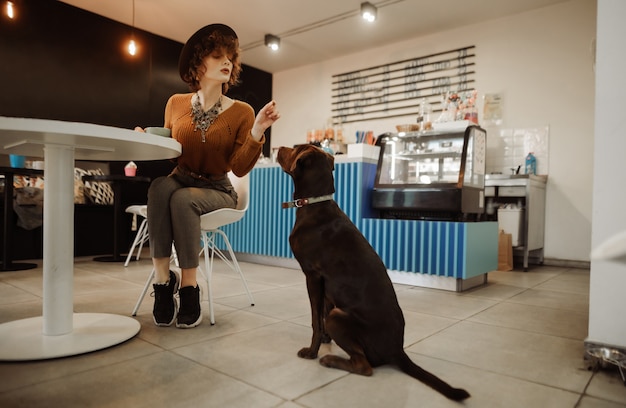 Beautiful girl in stylish clothes and hat playing with a dog in an animal friendly cafe
