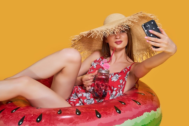 Beautiful girl in summer dress straw hat with inflatable ring shape of watermelon with fresh cocktai...