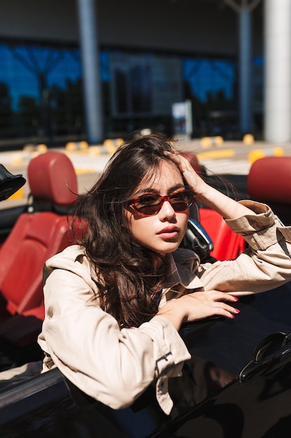 Beautiful girl in sunglasses and trench coat leaning on cabriolet car door while thoughtfully looking in camera with airport on background