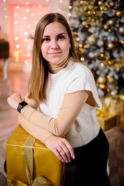 A beautiful girl in a sweater sits by a Christmas tree decorated with garlands. Next to it is a Christmas tree with boxes of gifts, decorated. New Year