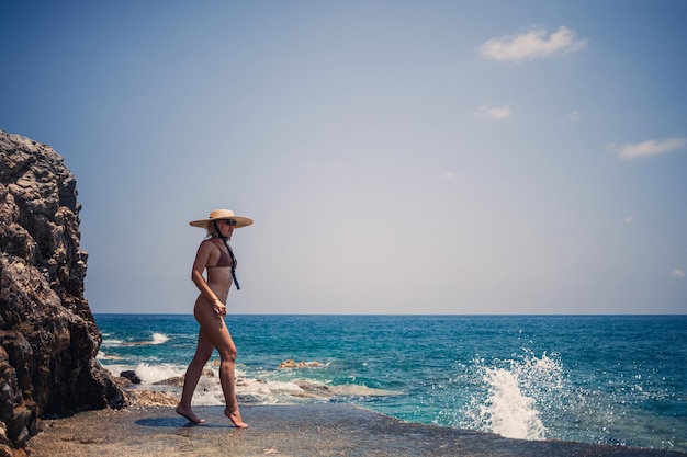 A beautiful girl in a swimsuit with a golden tan in a hat walks along the rocky coast near the blue sea Summer sunny day by the sea Vacation concept