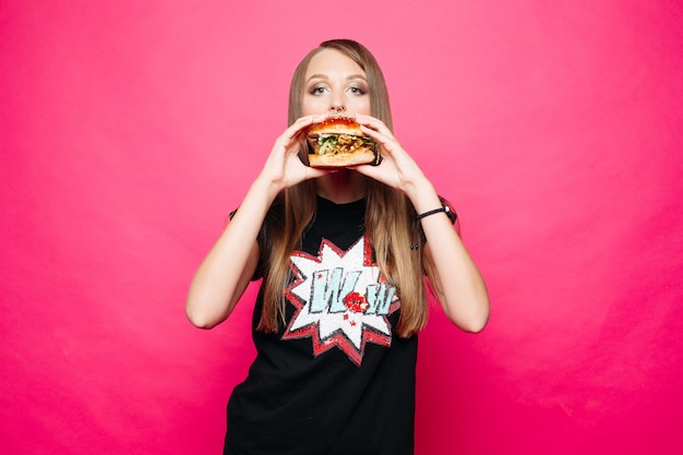 Bella ragazza in maglietta che mangia hamburger sopra il rosa luminoso