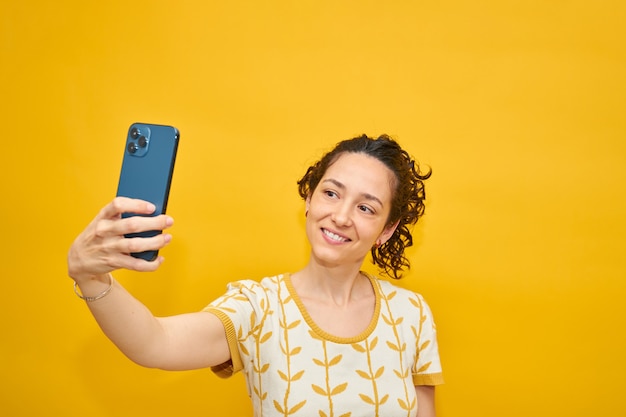 격리 된 노란색 배경에 selfie를 복용하는 아름 다운 소녀. 한 손으로 스마트 폰을 들고 카메라에 웃 고. 곱슬 머리.