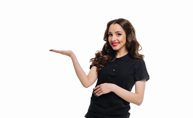 Beautiful girl waitress in black Tshirt and red lipstick stands on white background and holds her hand out with her palm smiling and looking at the camera The concept of service in the restaurant