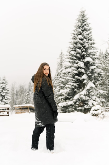 Beautiful girl walking in a snowfall. Young woman in black puffer jacket posing in the winter forest.Outdoor portrait of pretty brunette lady. Winter concept