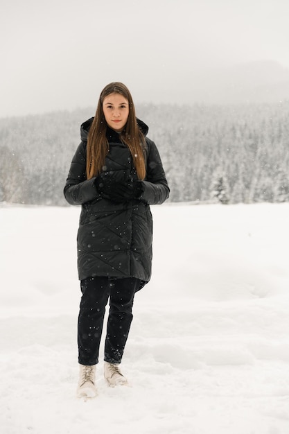 降雪の中を歩く美しい少女。冬の森でポーズをとる黒いフグジャケットの若い女性。かなりブルネットの女性の屋外の肖像画。冬のコンセプト